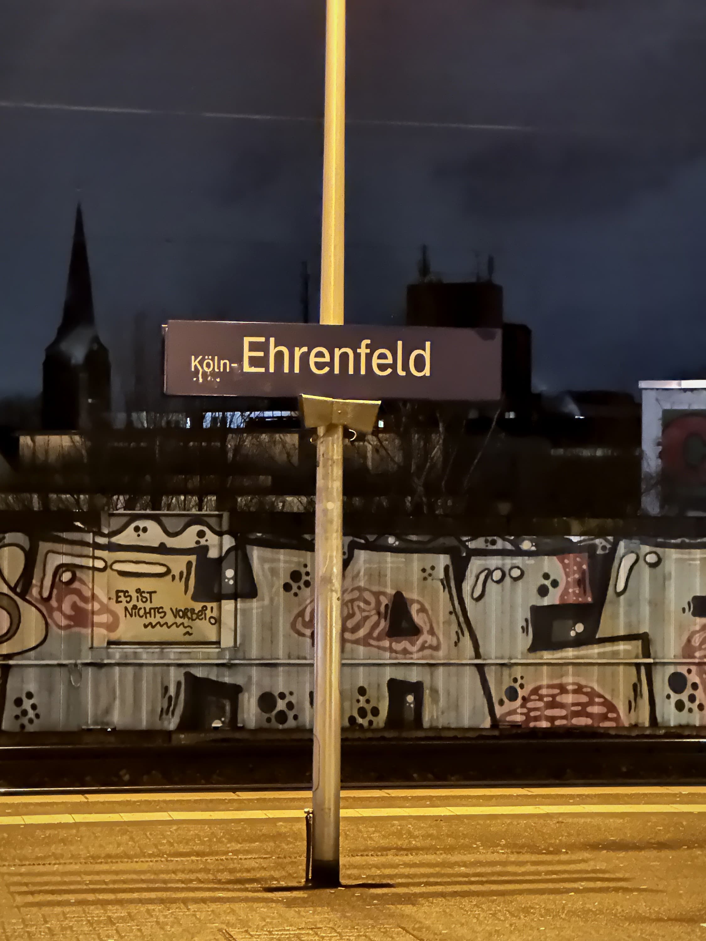 Bahnhofsschild Köln-Ehrenfeld vor einer Graffitiwand bei Nacht
